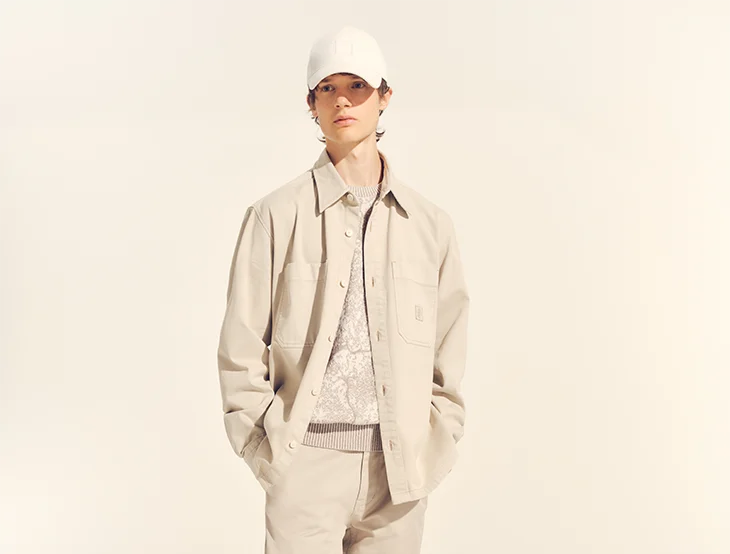 A male model posing in front of a light-colored background wearing sand-colored clothing (photo)