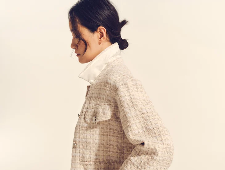 A female model posing in front of a light-colored background wearing sand-colored clothing (photo)