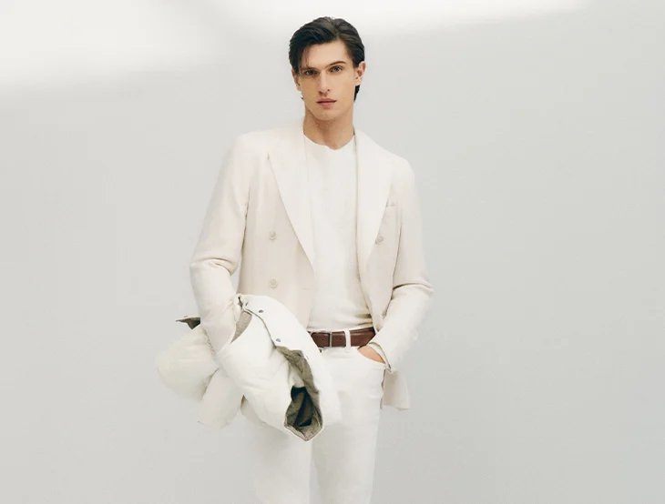 A male model posing in front of a light-colored background wearing white throusers and white jacket (photo)