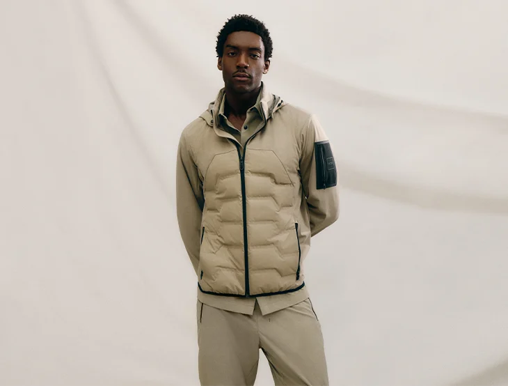 A male model posing in front of a light-colored background wearing sand-colored sports clothing (photo)