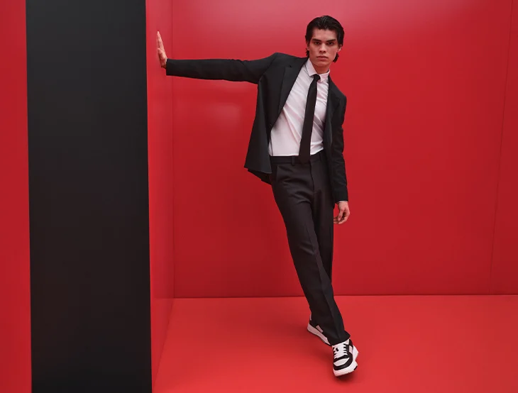 A male model posing in front of a red background wearing a black suit and sports shoes (photo)