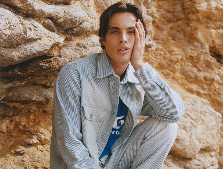 A male model posing in front of a rock wall wearing jeans clothing (photo)