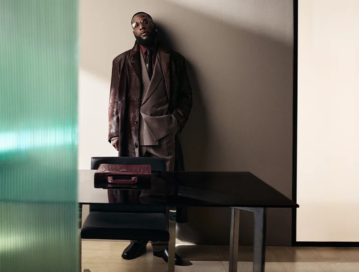 A male model posing in front of a sand-colored background with a table and a chair, wearing a brown suit (photo)